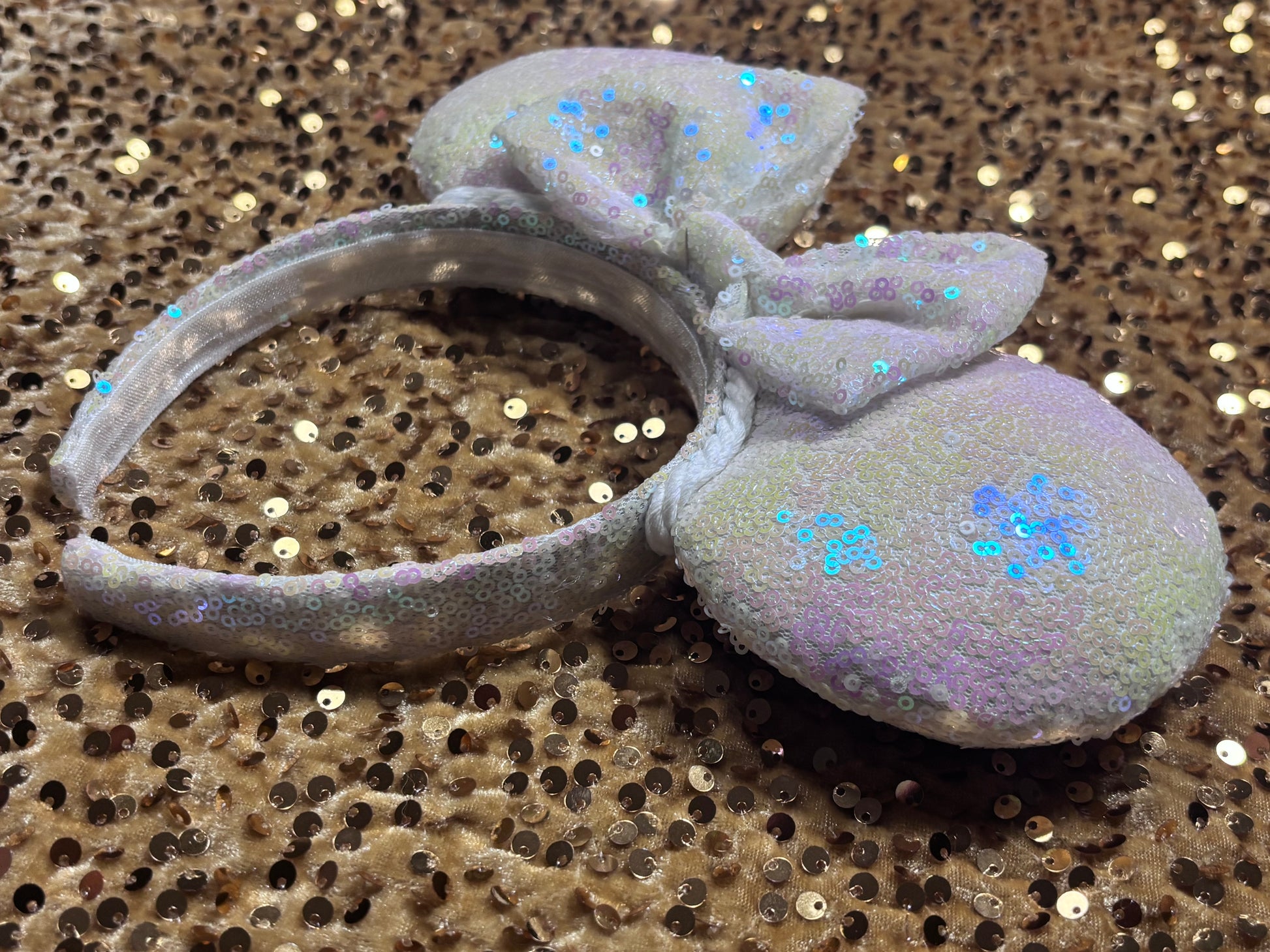 White Iridescent mouse ears with the fabric on both the front and back. The fabric is on the headband as well and is universal sized and fits most. The set has a matching iridescent bow sequin bow. Item is shown on a gold sequin background.