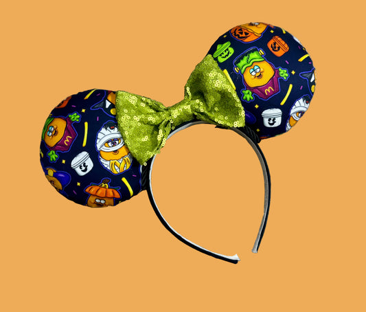 Halloween-themed ear headband featuring a navy blue fabric print with cartoon pumpkins, ghosts, and a green sequined bow, displayed on an orange background.