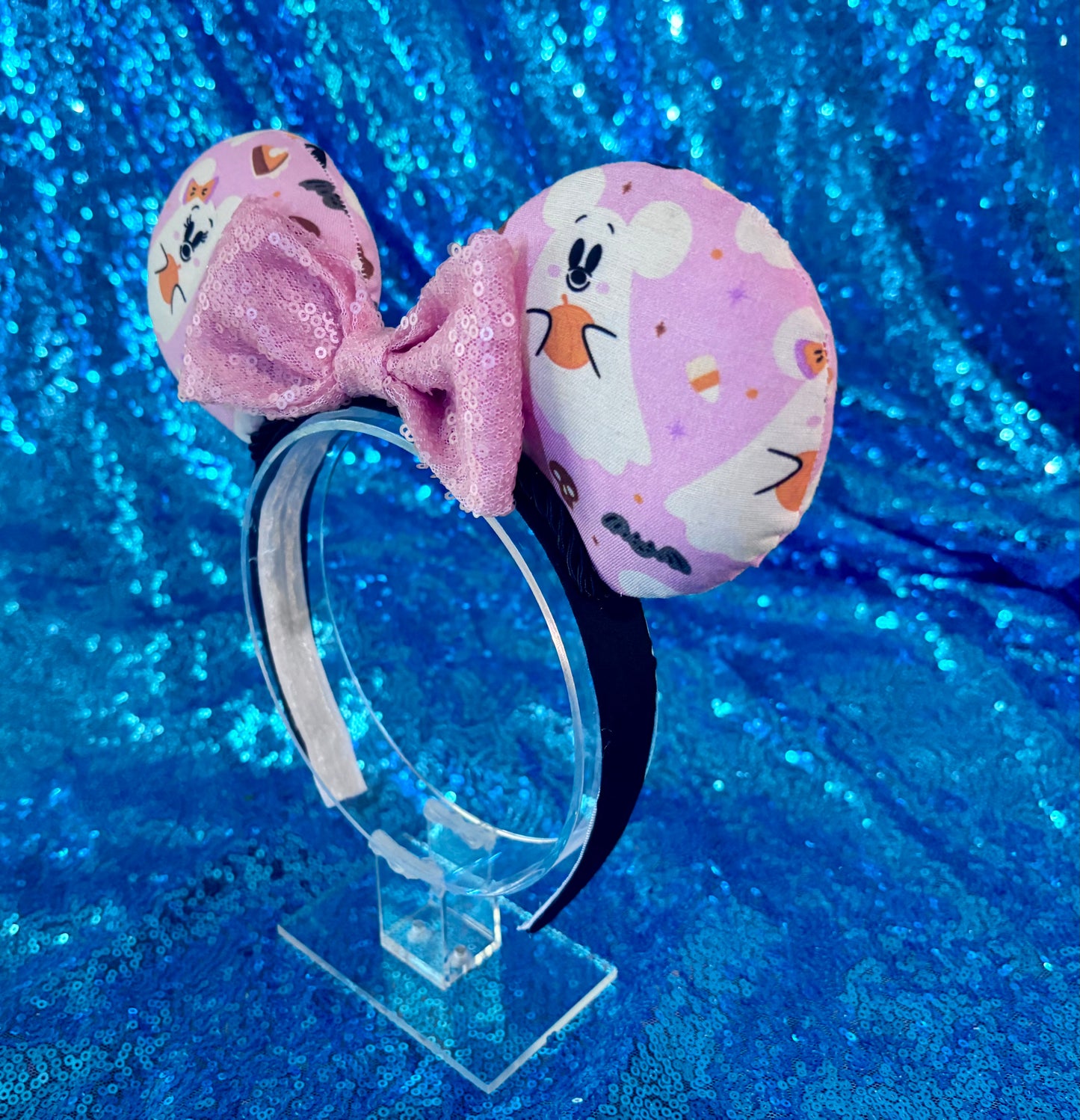 Side view of custom ear headband on a clear stand. Handmade and ready to ship Halloween Pink Ghosts Mouse ears with the fabric with mouse shaped ghosts on pink fabric is on the ears, front and back and a light pink sequin bow. The headband is a universal sized headband and has black poly cotton fabric on it.