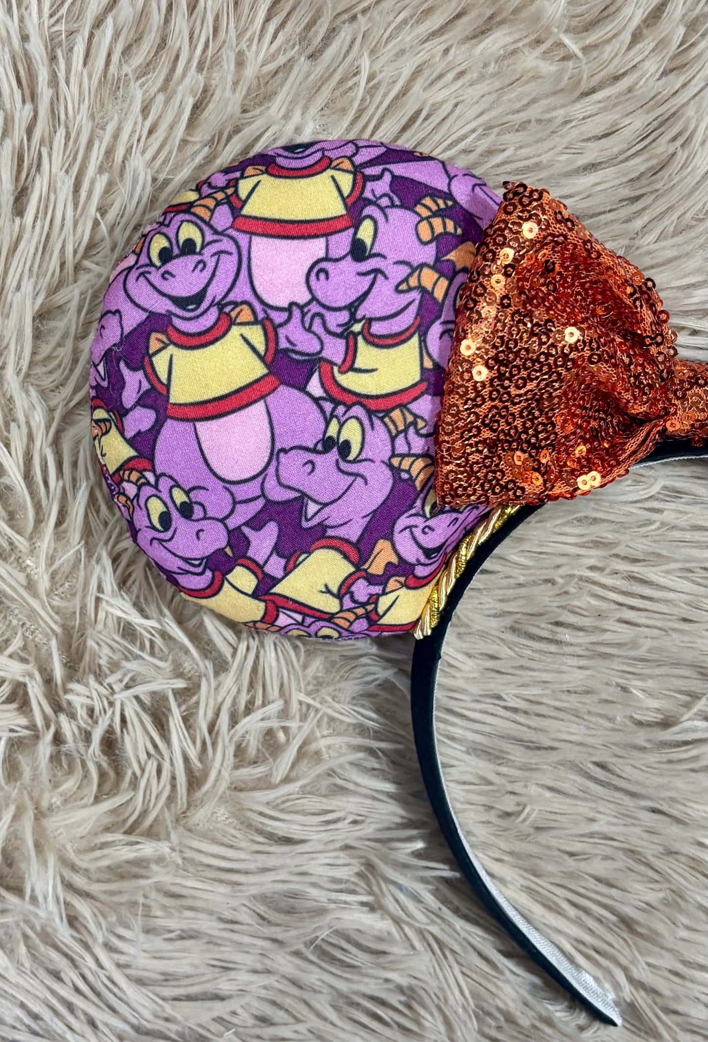 A close-up of a headband featuring a round fabric-covered ear in a purple cartoon dinosaur pattern with yellow and red accents, and a sequined orange bow on a shaggy beige rug.