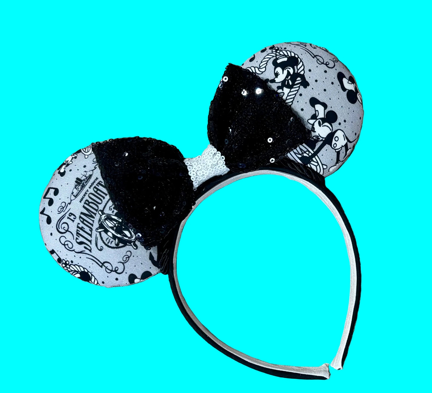 A pair of Mouse-themed ear headbands featuring a black and white patterned fabric with sequins, a black sequined bow with a white sequin middle, and a white headband, displayed on a beige faux fur background.