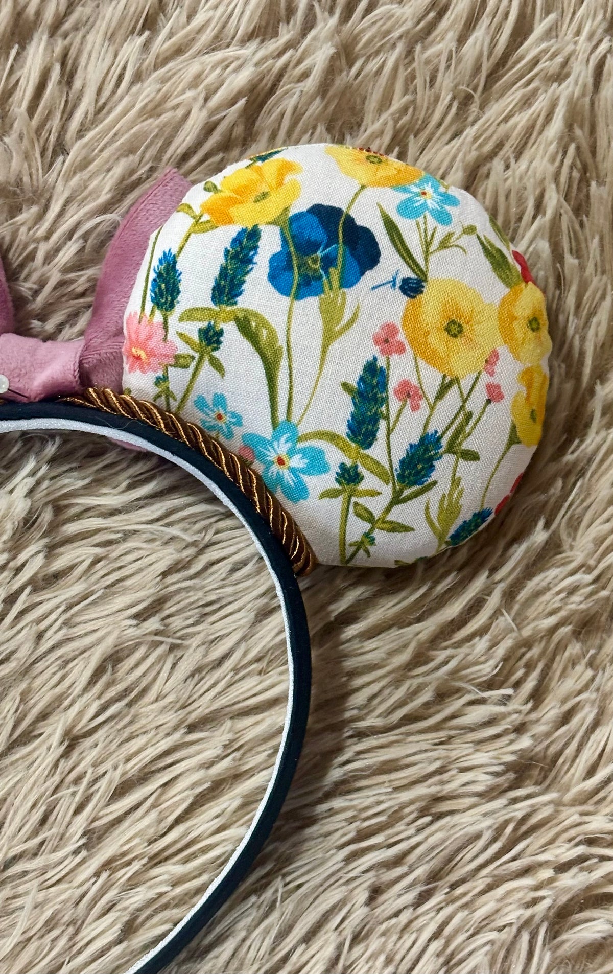 An ear headband accessory featuring a floral-themed print on the ears and a pink velvet bow in the center with copper colored twisted trim along the inner ears.