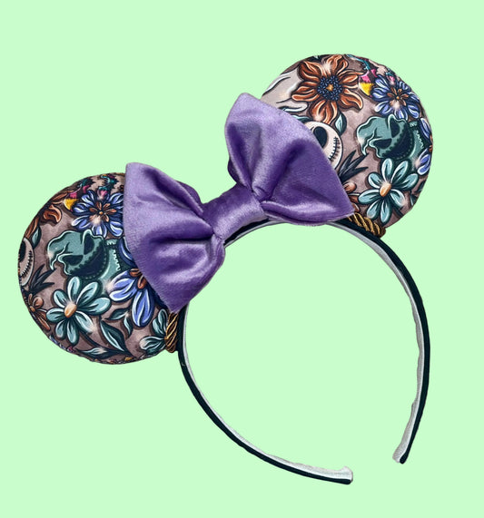 A pair of ear headband accessories with a purple velvet bow in the center and floral-patterned ears on a green background.