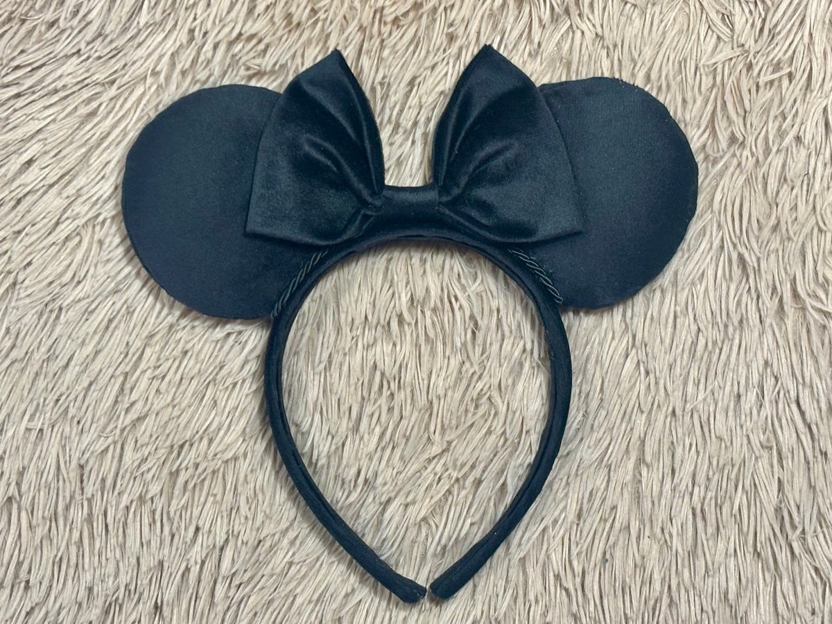 Black velvet ears headband with a large bow, laid flat on a beige faux fur surface.