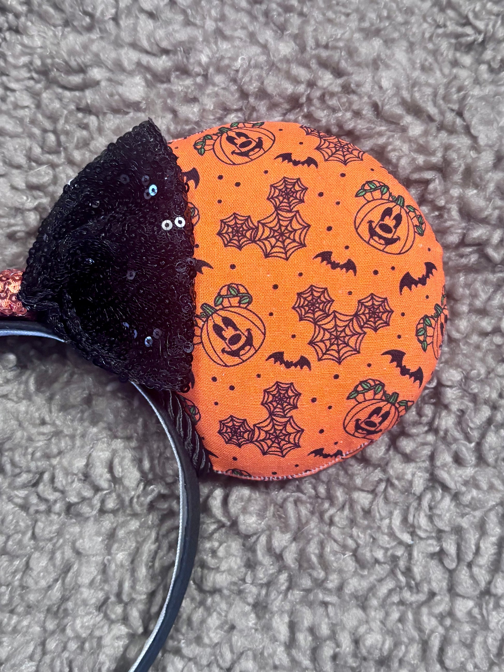 Halloween mouse ears with the fabric on both the front and back. It's orange with bats, mouse shaped pumpkins and spider webs. The headband is black universal sized and fits most. The set has a black with orange middle sequin bow. Item is shown on a brown furry background.