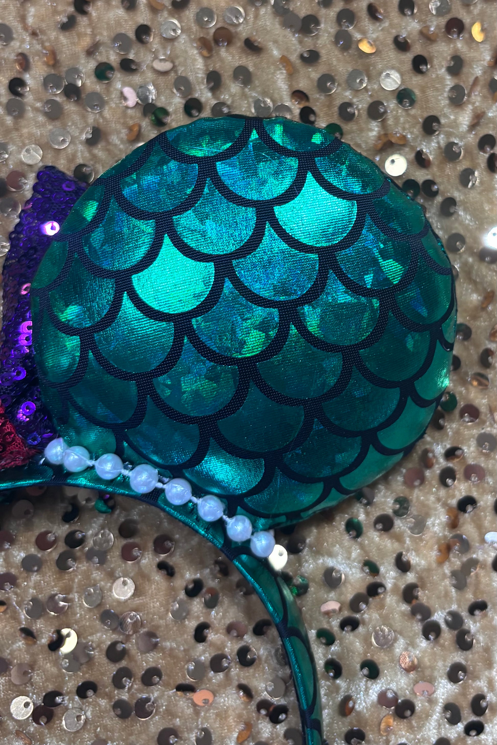 Mermaid-themed headband with teal scales and a purple sequin bow with a red middle and a dinglehopper middle piece. Pearl garland trim along inner ears m on a matching mermaid scales headband. Other back of ear close up.