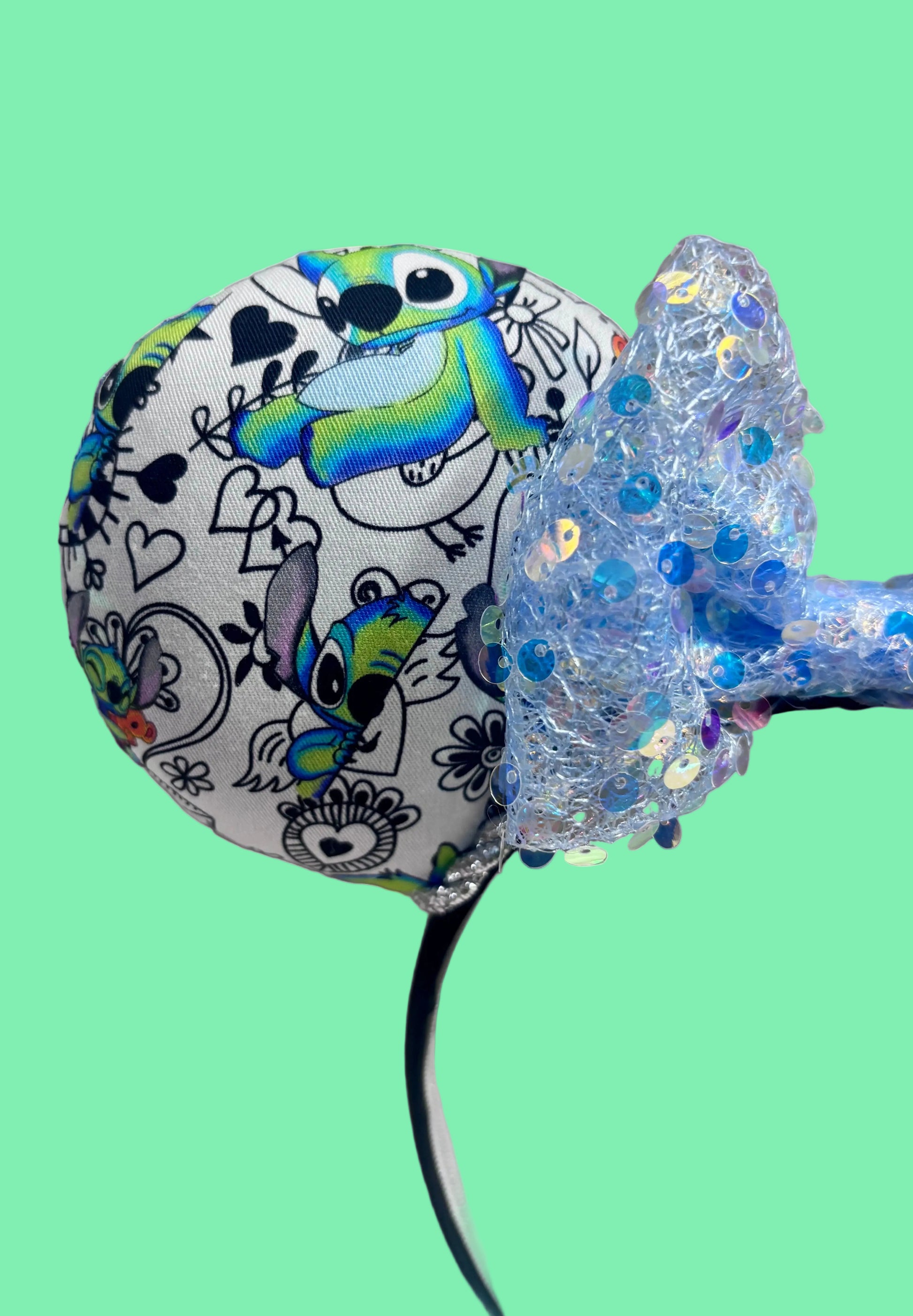 Close up view of front mouse ear. Handmade and ready to ship Mouse ears with Stitch cartoon character design on the fabric with silver twisted trim on a black universal sized headband and a blue sequin bow.