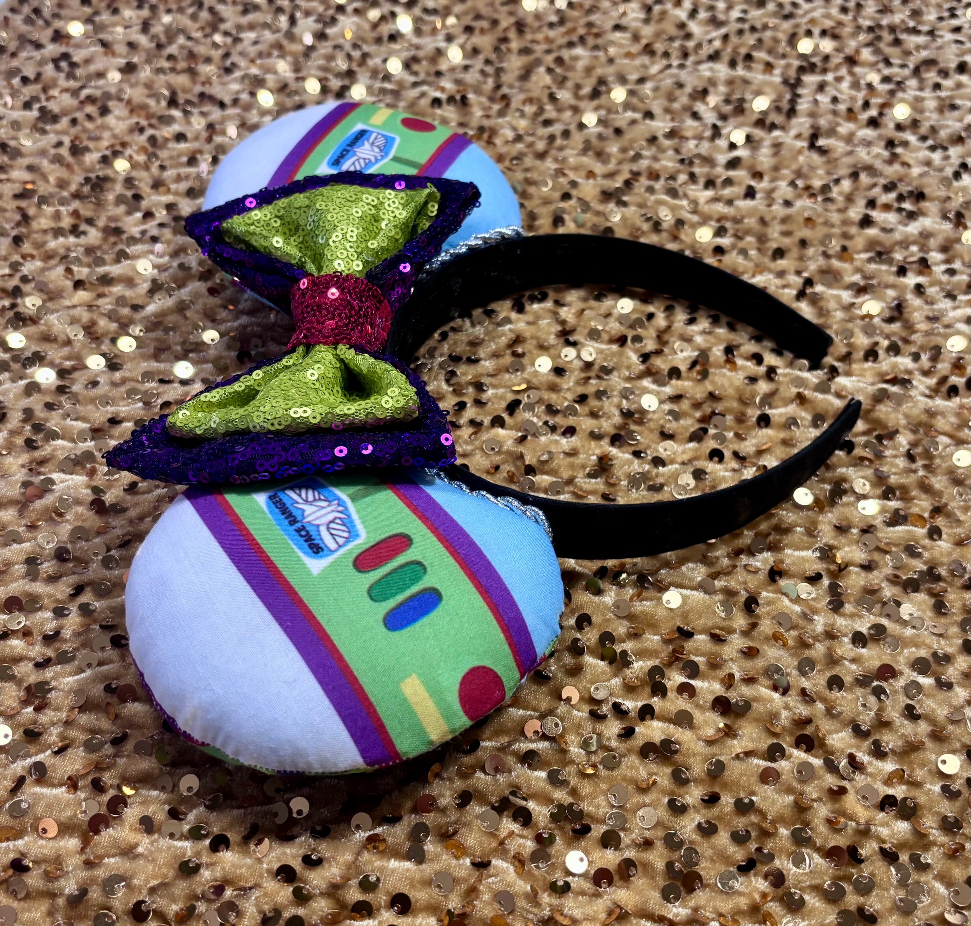 Space Ranger Mouse Ear headband with new improved bow. Handmade and ready to ship with Space ranger fabric on both the front and the back of mouse ears. The set has a silver twisted trim along the inner ears and a new sequin bow that is purple, lime green and red in sequin. Ears are on a black poly cotton universal sized headband. Side view of ears and headband.