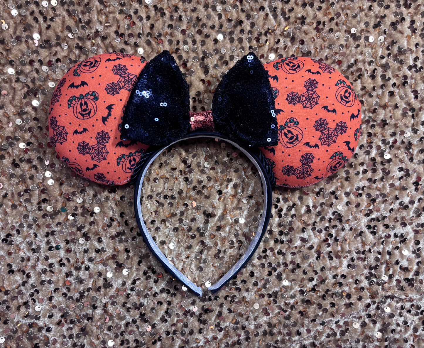Halloween mouse ears with the fabric on both the front and back. It's orange with bats, mouse shaped pumpkins and spider webs. The headband is black universal sized and fits most. The set has a black with orange middle sequin bow. Item is shown on a gold sequin background.