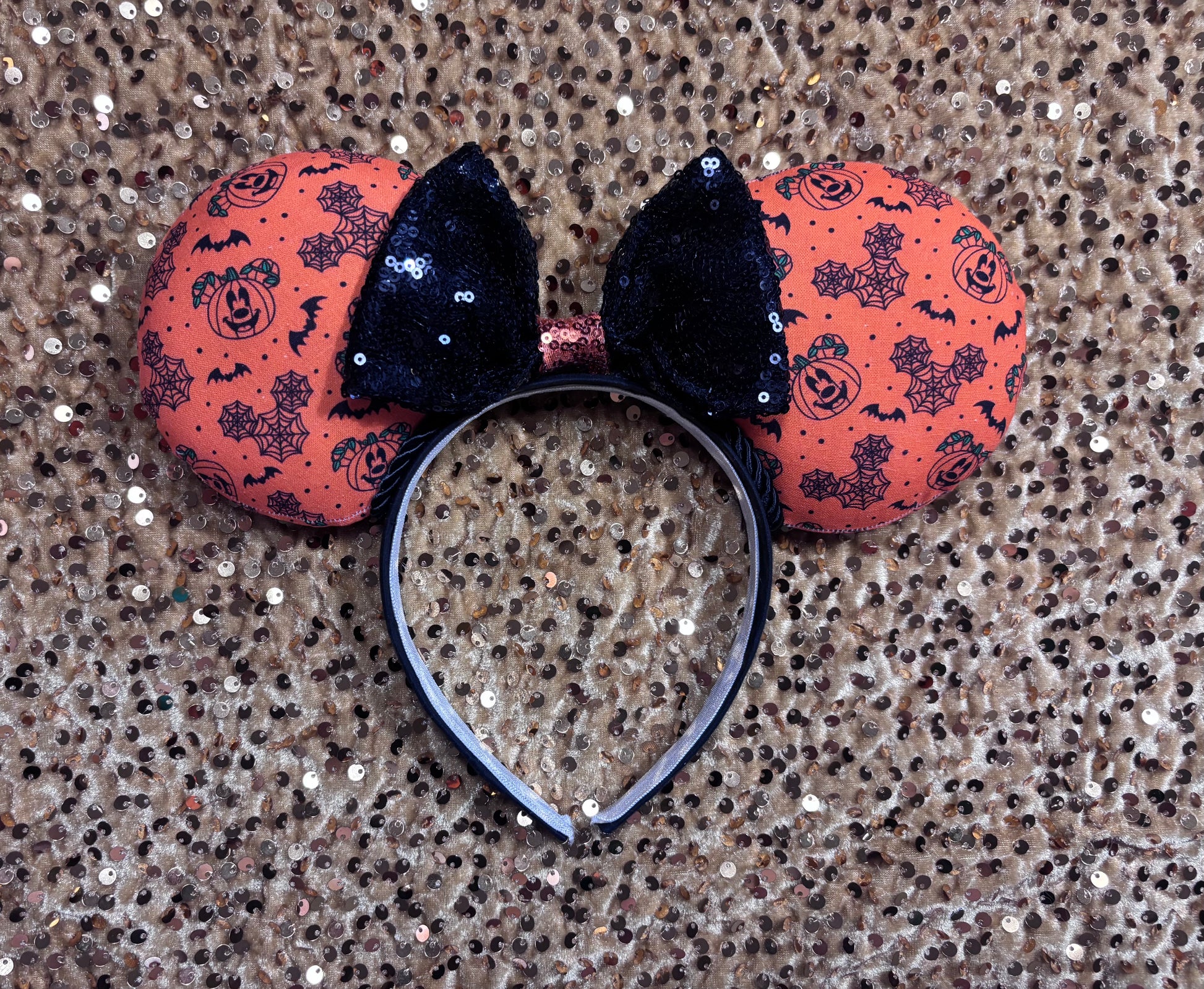 Halloween mouse ears with the fabric on both the front and back. It's orange with bats, mouse shaped pumpkins and spider webs. The headband is black universal sized and fits most. The set has a black with orange middle sequin bow. Item is shown on a gold sequin background.