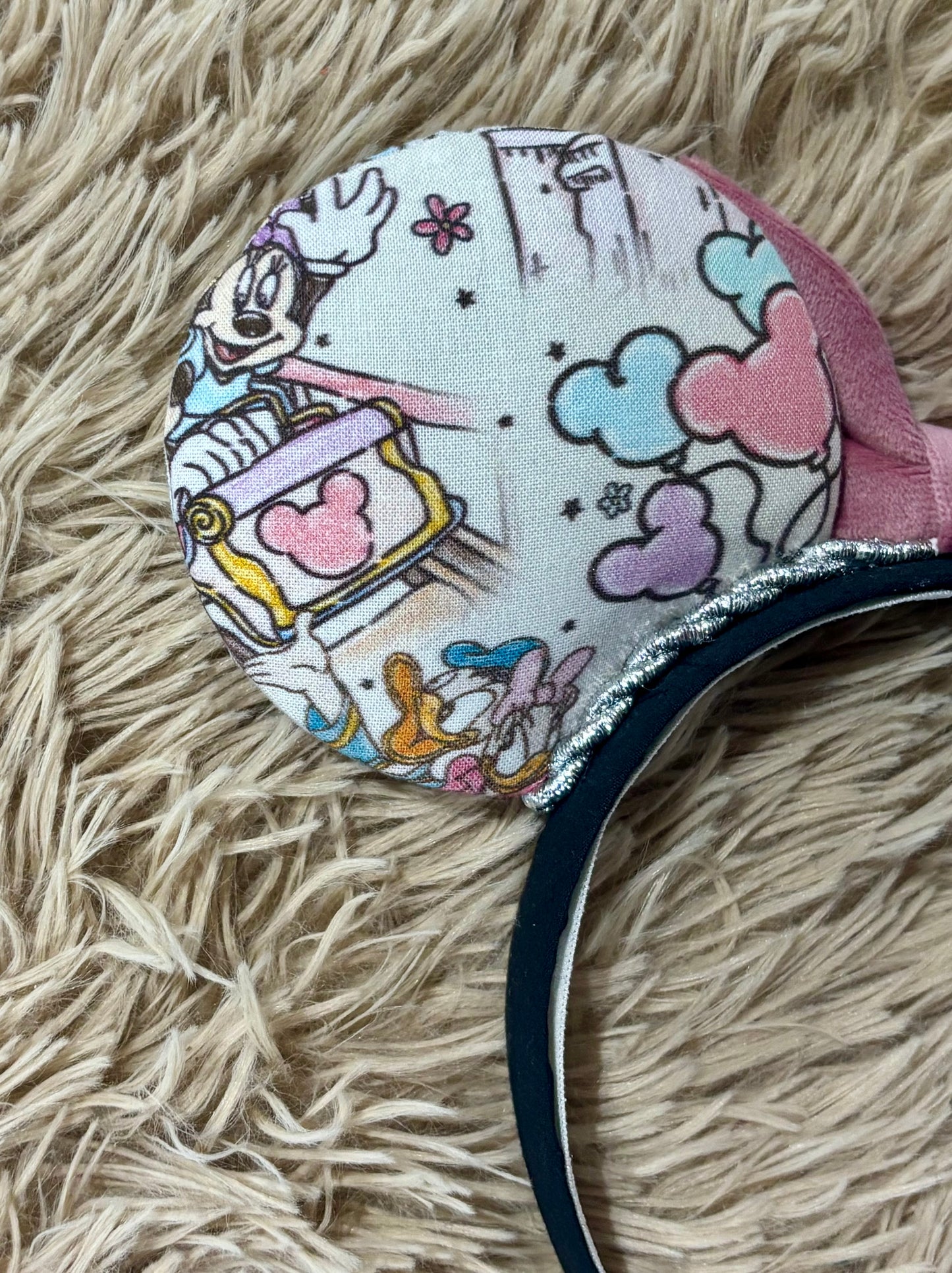 A close-up of a round fabric-covered headband or accessory featuring a multicolor cartoon print with hearts, stars, and character artwork, attached to a black band with a silver glitter trim, resting on a beige shag rug.