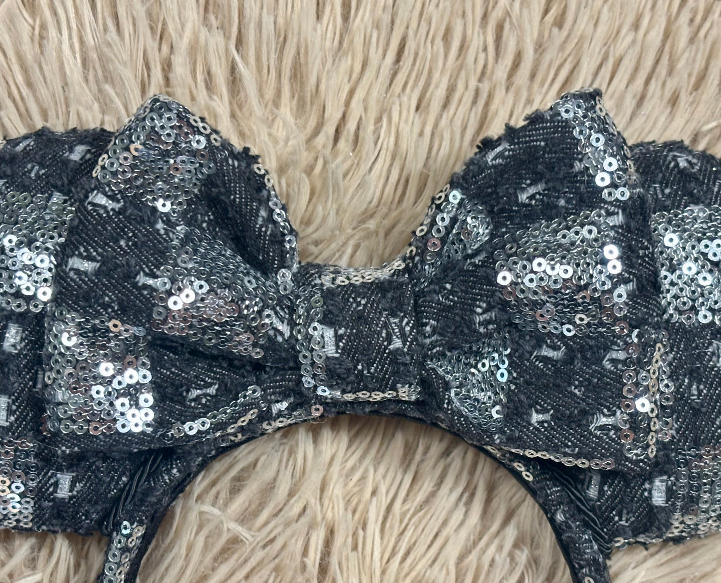 A close-up of a large bow headband covered in black sequins with silver reflective accents, laid flat on a beige faux-fur surface.