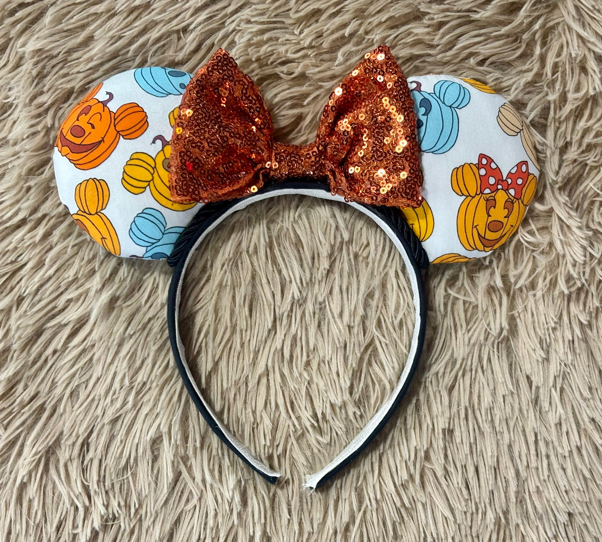 A Pumpkin-themed ear headband featuring a light white background with orange, yellow, and blue pumpkins, a large sequined orange bow, and a black headband.