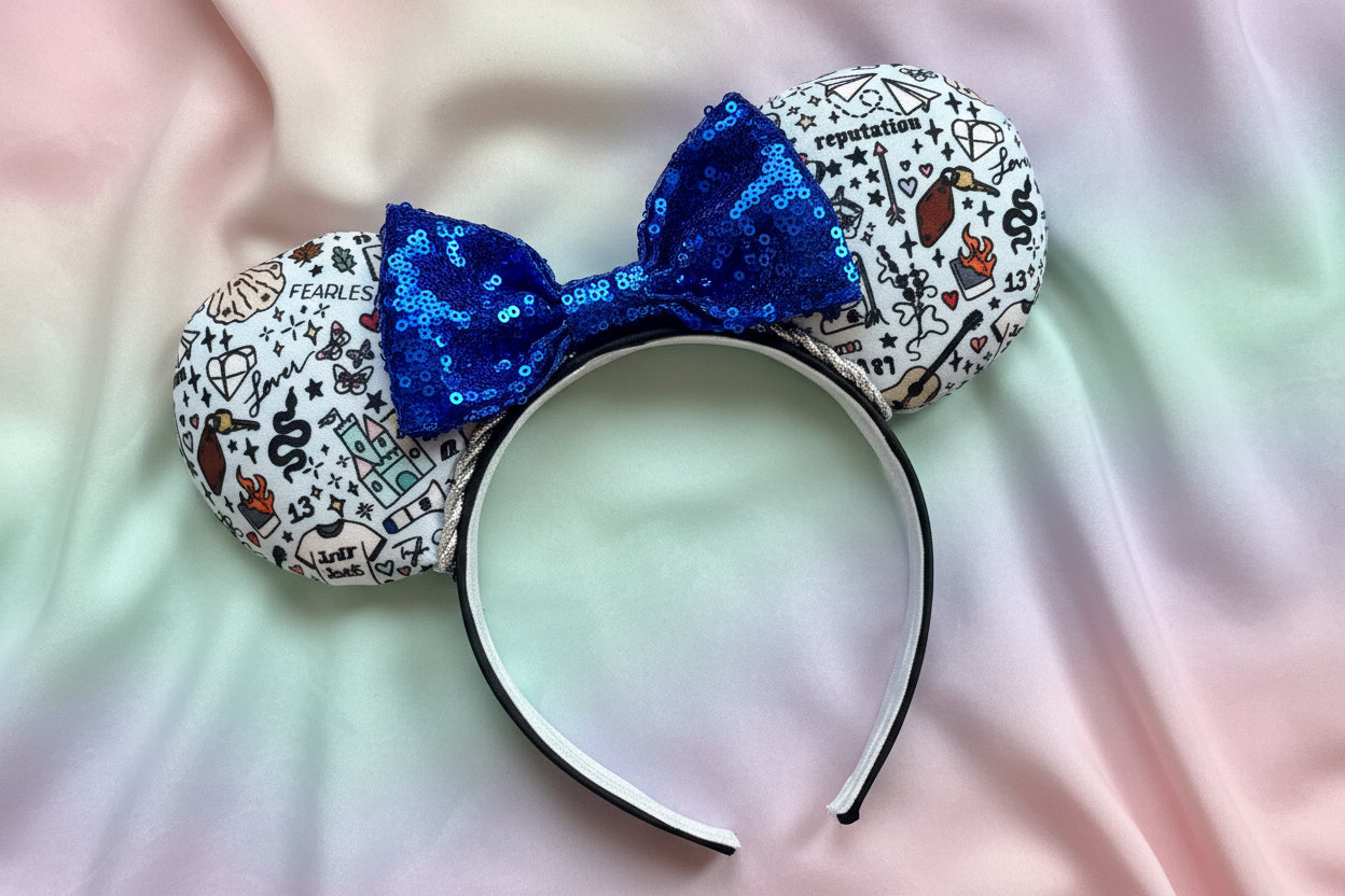 Swiftie Doodles from various albums in cartoon print on the mouse ears with a blue sequin bow on a black headband. Shown on a colorful fabric background