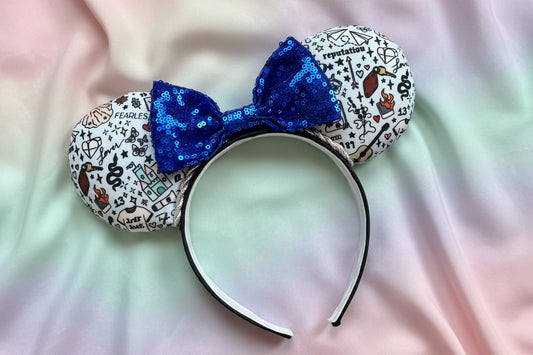 Swiftie Doodles from various albums in cartoon print on the mouse ears with a blue sequin bow on a black headband. Shown on a colorful fabric background