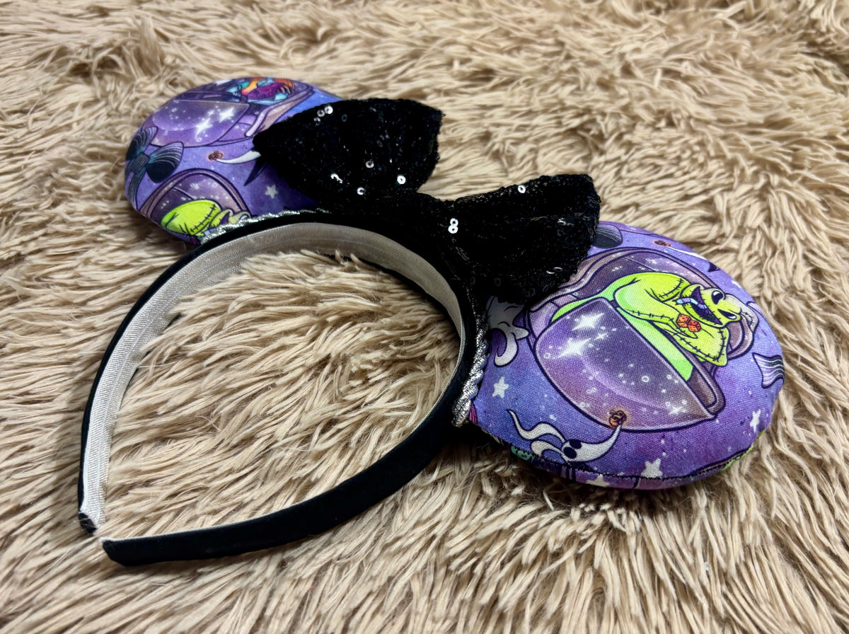 An ear headband accessory with a black sequined bow, featuring a purple HM and NBC themed print with a cartoon character in a buggy.