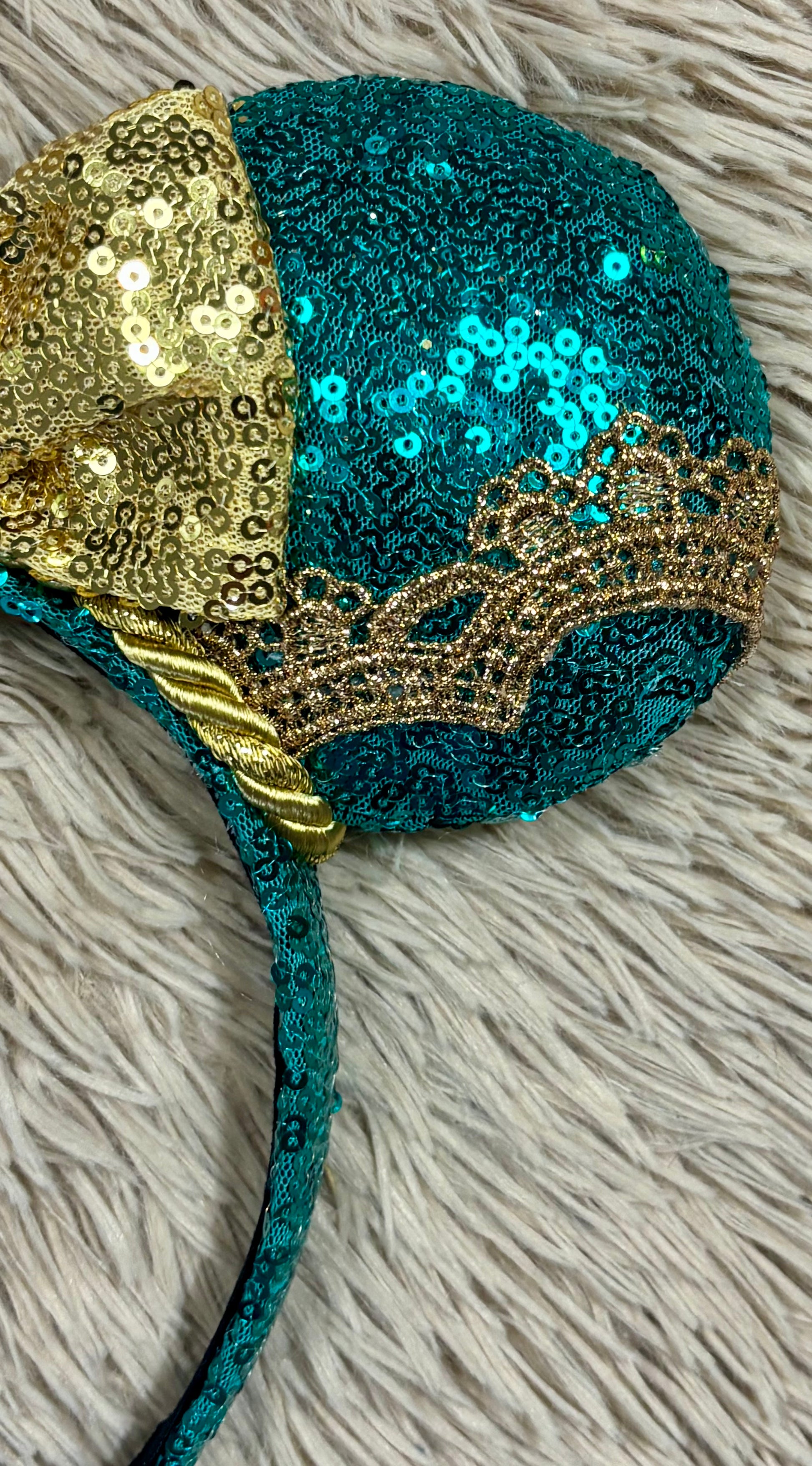 A close-up of a teal sequined headband with a large gold sequin bow and a gold tiara design on the ear cup, resting on a white faux fur surface.