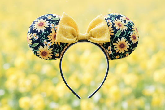 Yellow Floral minnie ears with a yellow sequin bow and gold twisted trim on a black headband. Shown on a flower field background
