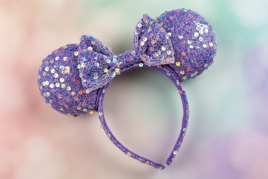 Purple sequin mouse ears on the ears, headband and bow. Purple twisted trim along inner ears. Shown on a blurred multicolored background