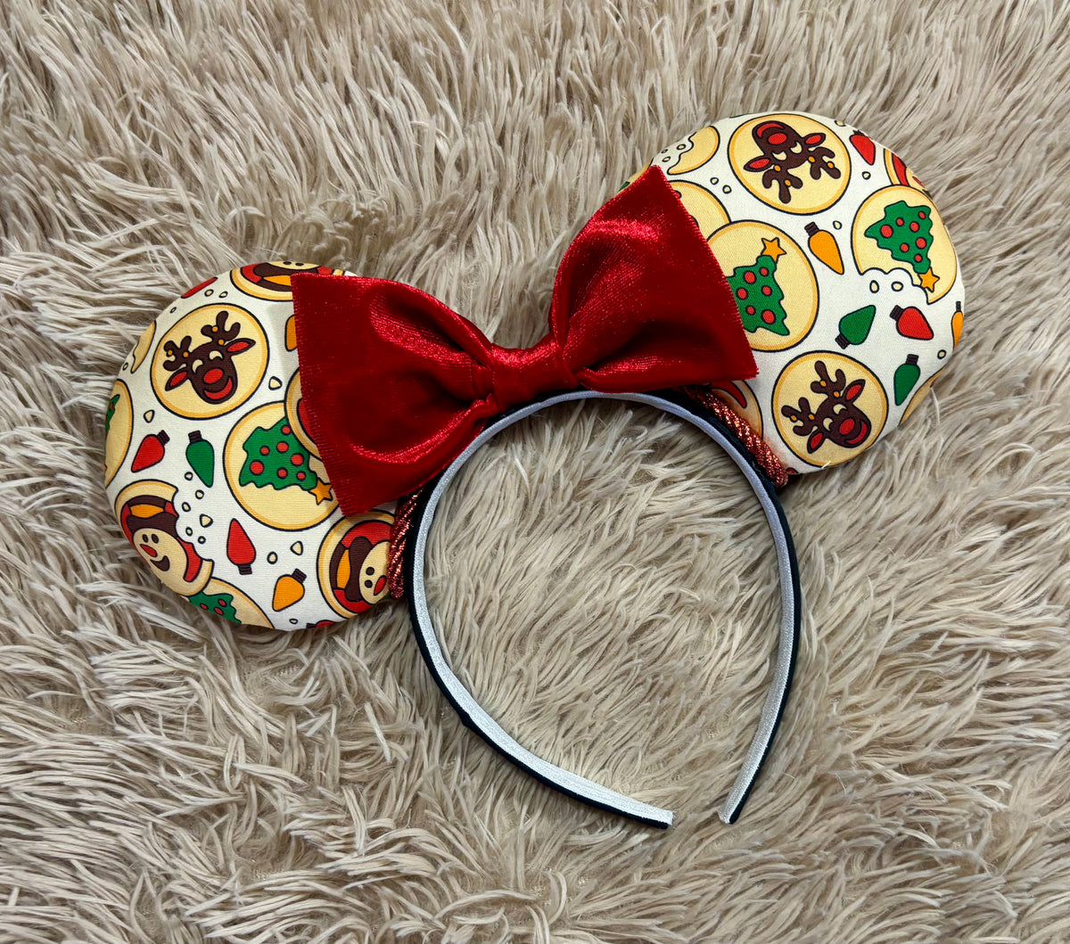 An ear headband featuring a festive Christmas print with red velvet bows, displayed on a beige faux fur background.
