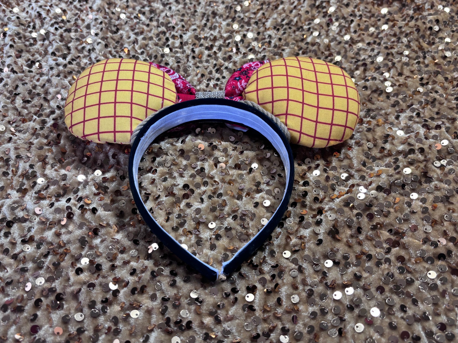 Handmade and ready to ship. Sheriff Woody Inspired mouse ears with the fabric on both the front and back that looks like his shirt. The fabric on the headband is black and is universal sized and fits most. The set has a bandana and cow print layered bow with brown trim along the inner ears to look like rope. Item is shown on a  gold background.