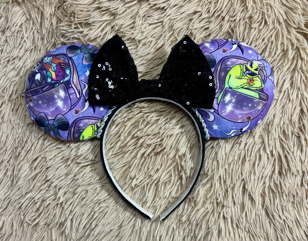 An ear headband accessory with a black sequined bow, featuring a purple HM and NBC themed print with a cartoon character in a buggy.