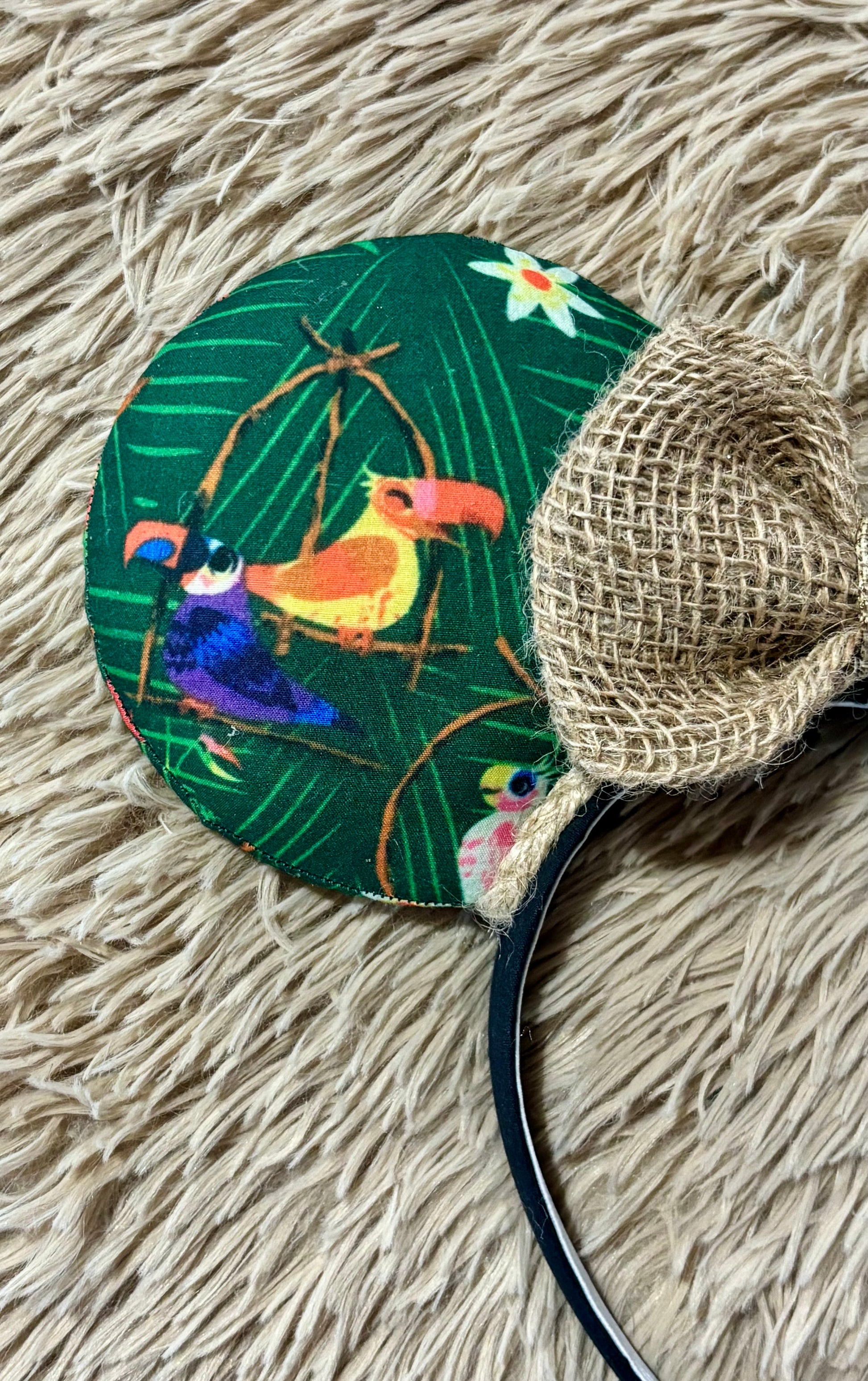 A close-up of a round fabric headband featuring a tropical toucan design on a dark green background, with a woven burlap-look ear accent and a black headband band visible at the bottom right, laid on a beige faux-fur surface.
