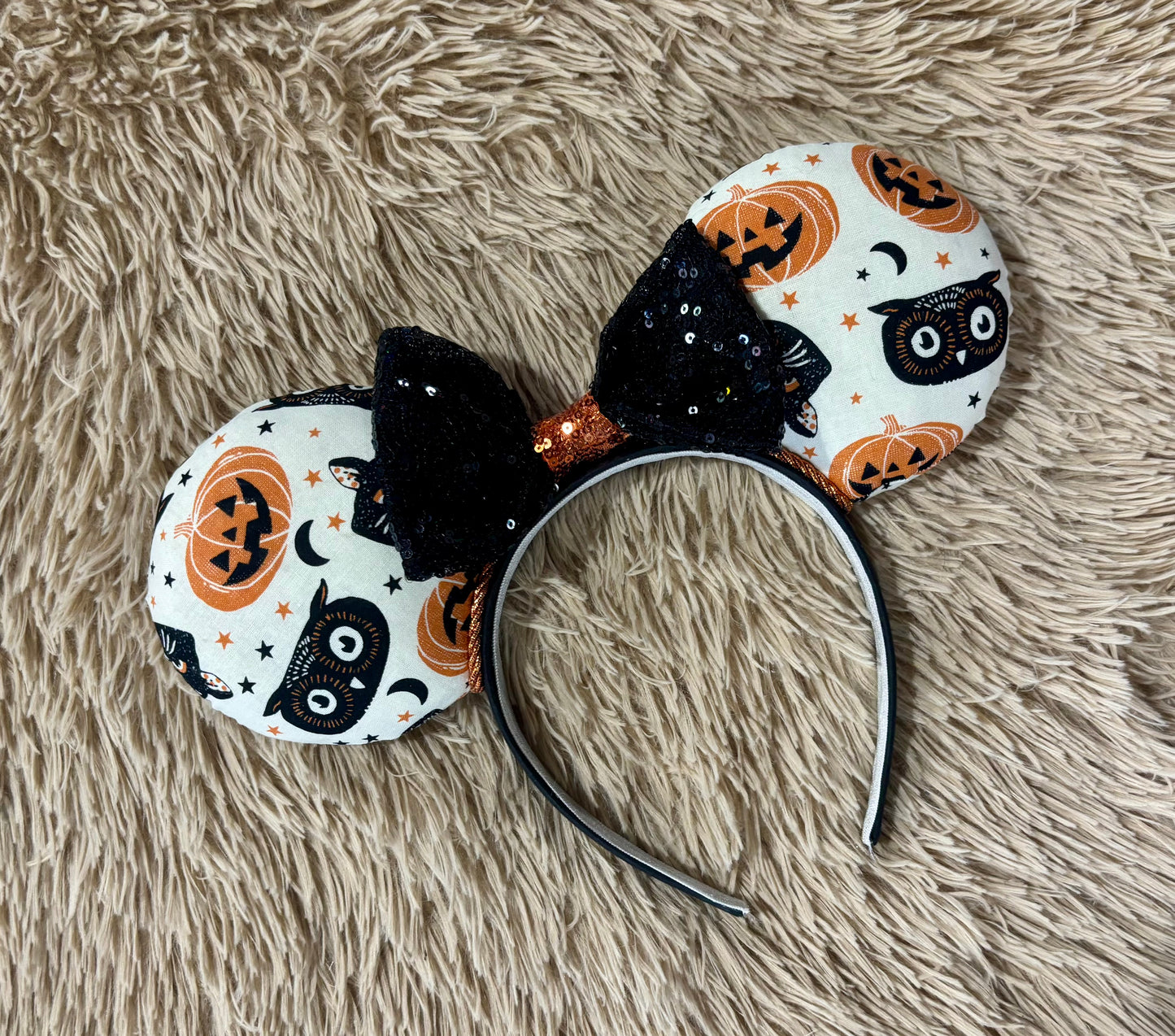 A Halloween-themed ear headband featuring a white fabric print with black cats, orange pumpkins, and stars, accented by a large black sequined bow with an orange glittery center.