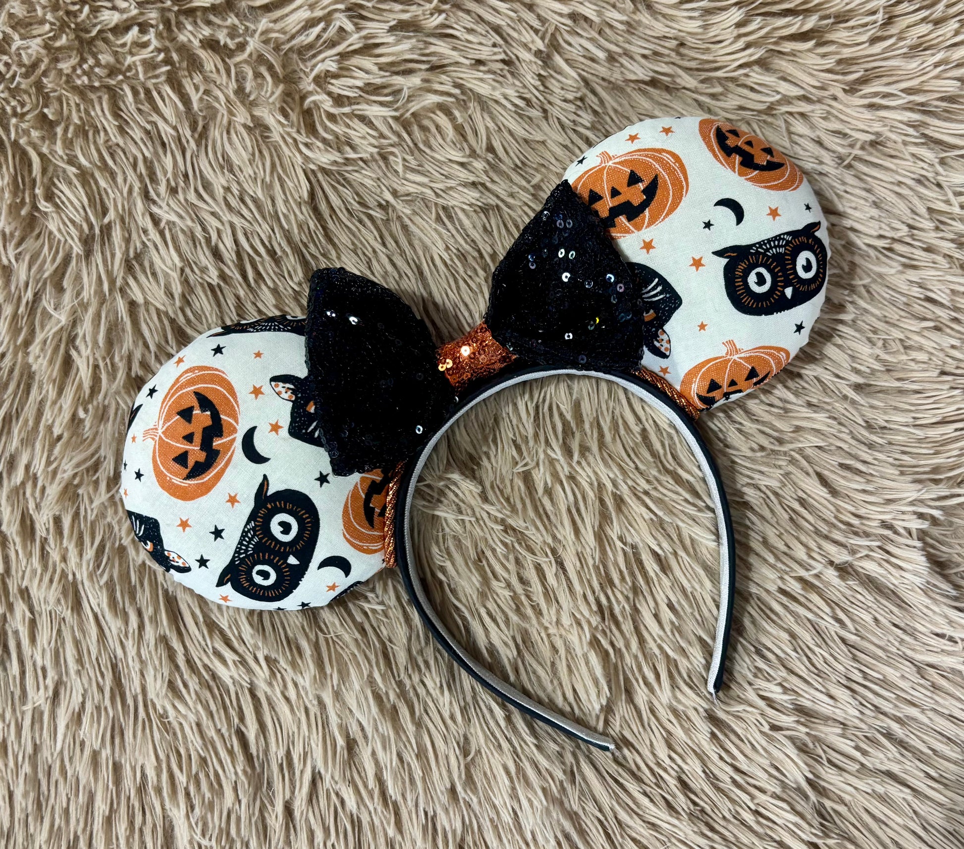 A Halloween-themed ear headband featuring a white fabric print with black cats, orange pumpkins, and stars, accented by a large black sequined bow with an orange glittery center.