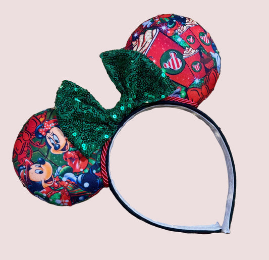 A festive ear headband featuring holiday-themed printed fabric on the ears and a large green sequined bow, shown against a solid pale pink background.