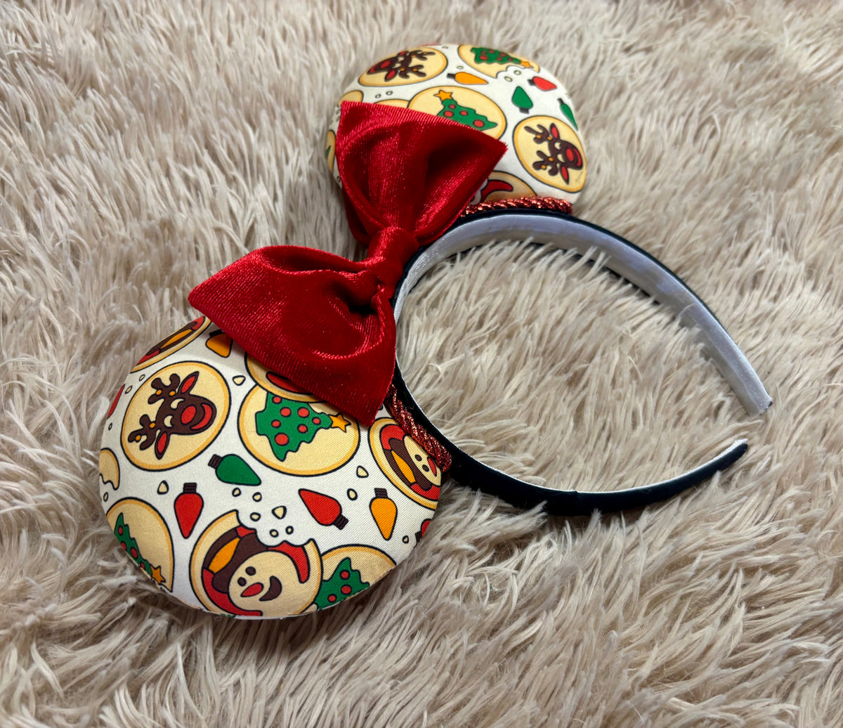 An ear headband featuring a festive Christmas print with red velvet bows, displayed on a beige faux fur background.