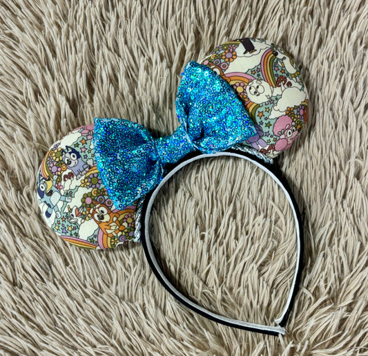 An ear headband with a colorful cartoon print and a large glittery blue bow, displayed on a beige faux-fur surface.