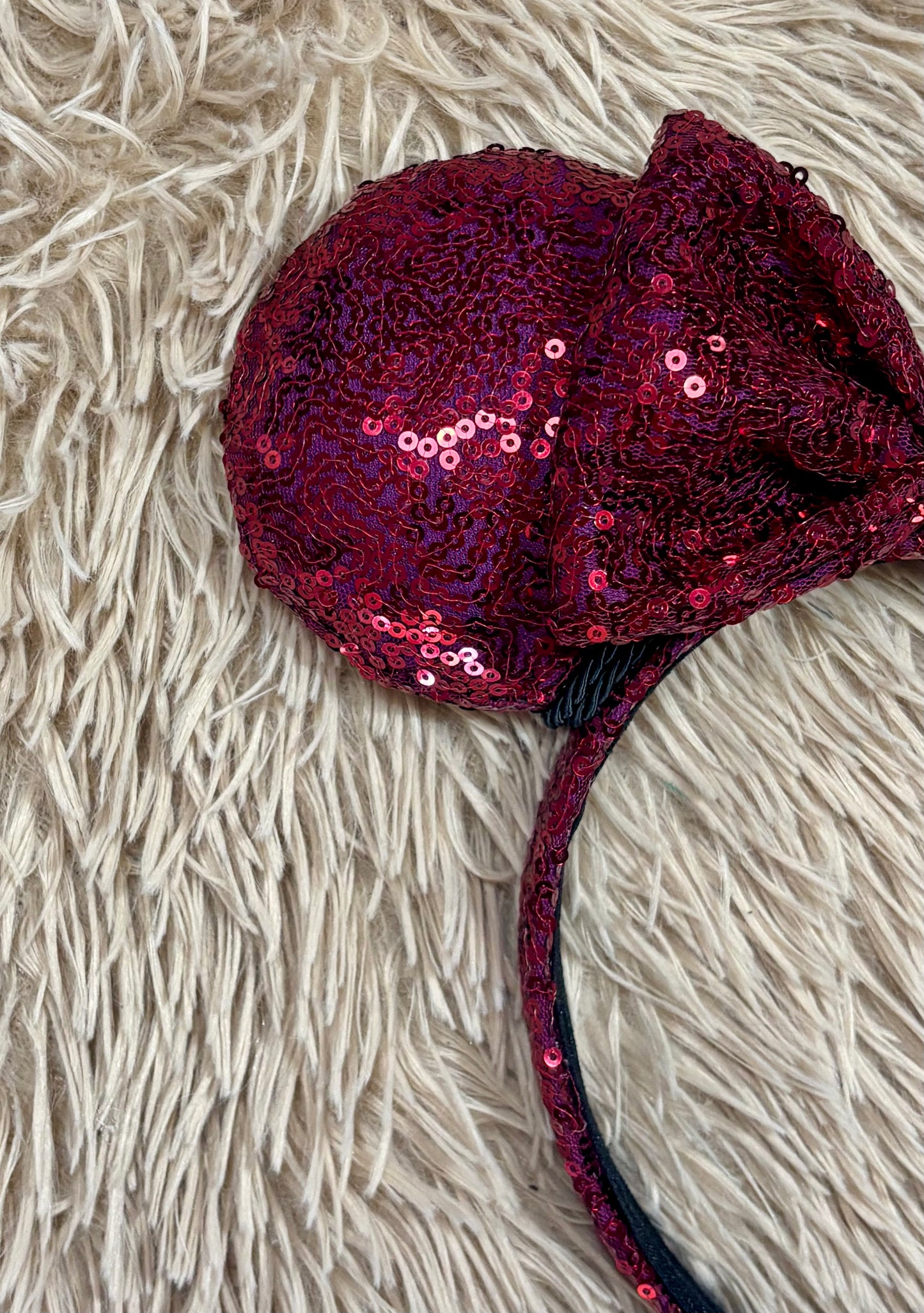 A close-up of a dark red sequined headband with a large bow on a textured beige faux fur background.