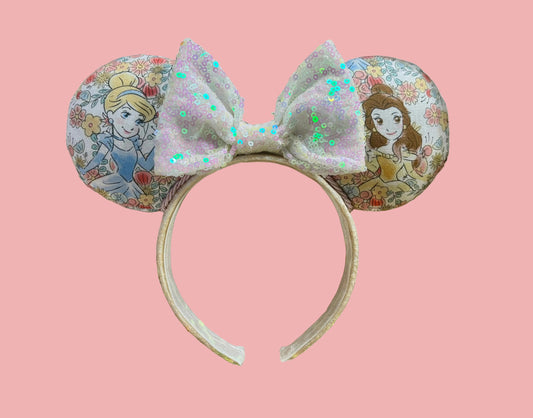 Handmade and ready to ship Pretty Princess Mouse ears with Cinderella and Belle shown on the fabric with a white iridescent bow. The headband has a white shimmery fabric and is a universal sized headband.