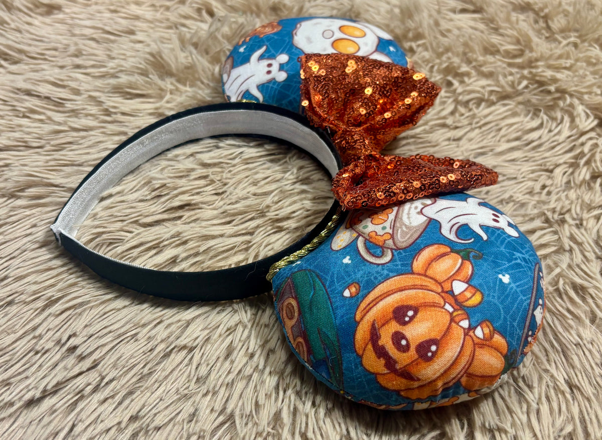 Halloween-themed headband featuring round blue ears with a pumpkin and ghost print, a glittery orange sequin bow, and a black elastic band, displayed on a beige faux fur background.