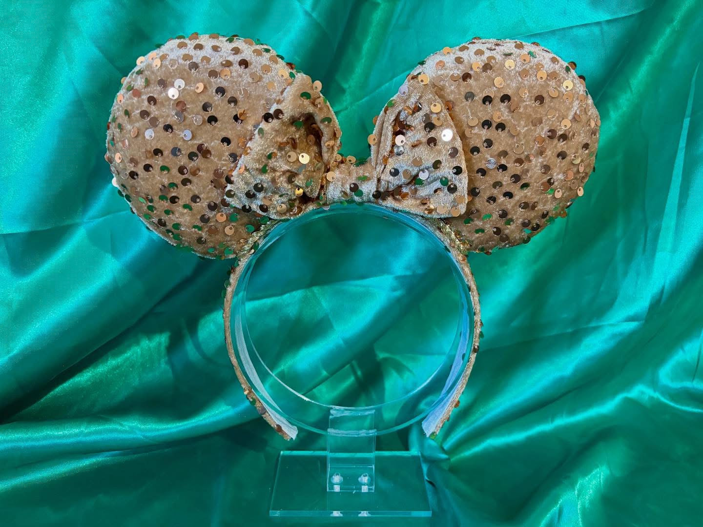 Gold velvet sequin mouse ears on the headband, bow and mouse ears. Shown on a clear stand against a teal fabric background