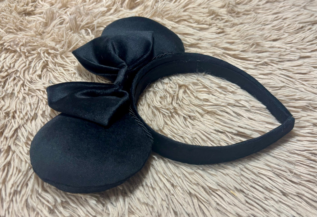 Black velvet ears headband with a large bow, displayed on a beige faux fur background.