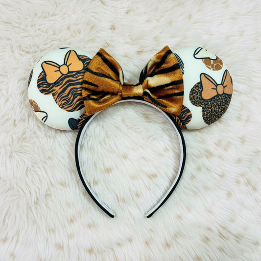 A ear headband featuring a white fabric with tiger, leopard, and cheetah print patterns, accented by a large metallic gold and black striped bow on a black headband, displayed on a beige faux fur background.