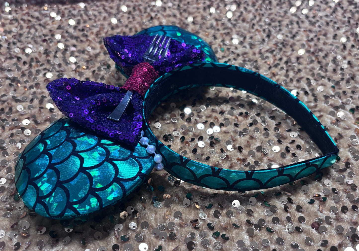 side view of headband. Mermaid-themed headband with teal scales and a purple sequin bow with a red middle and a dinglehopper middle piece. Pearl garland trim along inner ears m on a matching mermaid scales headband.