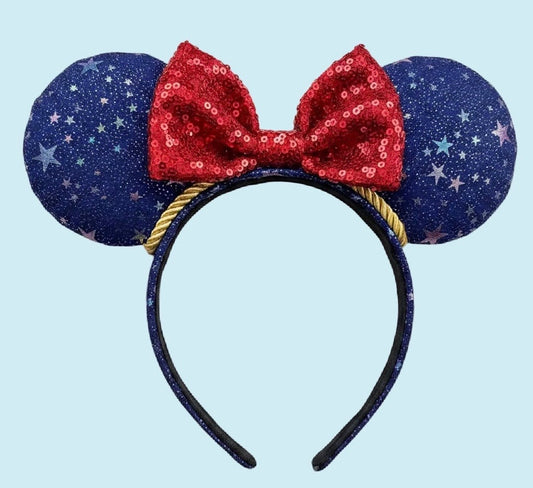 An ear headband featuring navy blue fabric ears with white star patterns, a red sequined bow, and a gold rope accent on a light blue background.