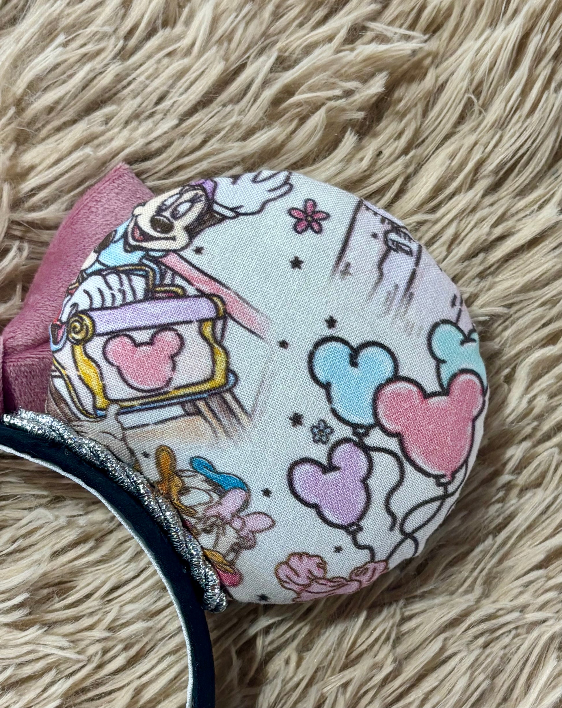 A pair of handmade ear headbands with a large dusty pink velvet bow, featuring printed park themed patterns on the ears and resting on a beige faux fur surface. Close up of back ear.