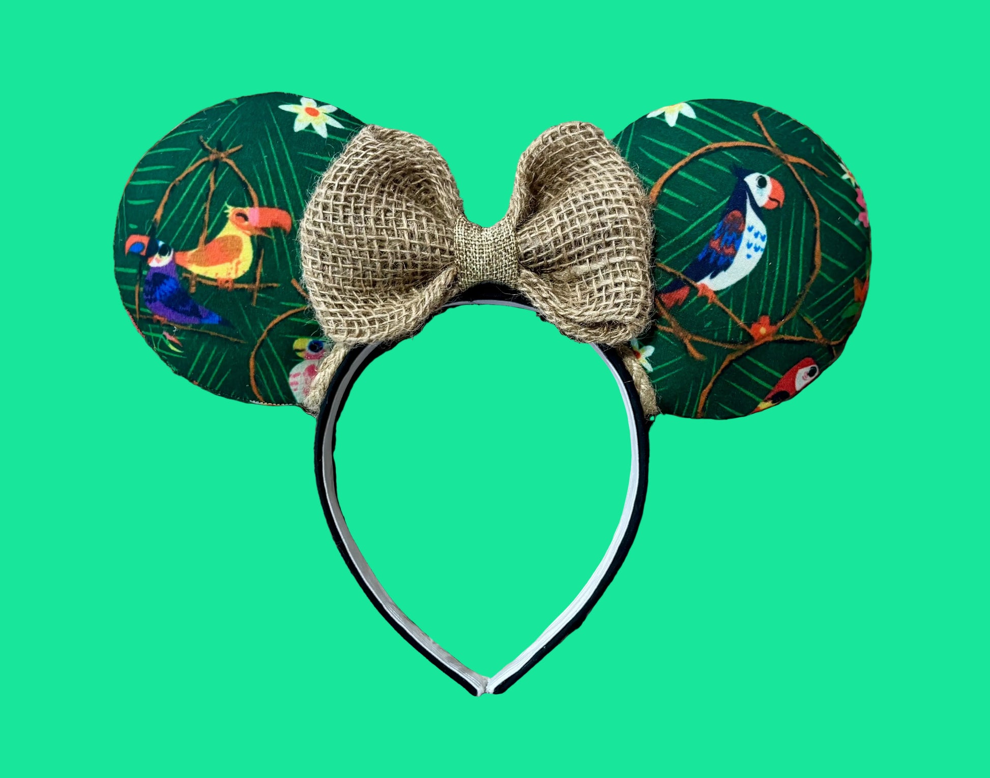A pair of ears featuring green tropical fabric with toucan and bird prints, a large burlap bow in the center, and a black headband.