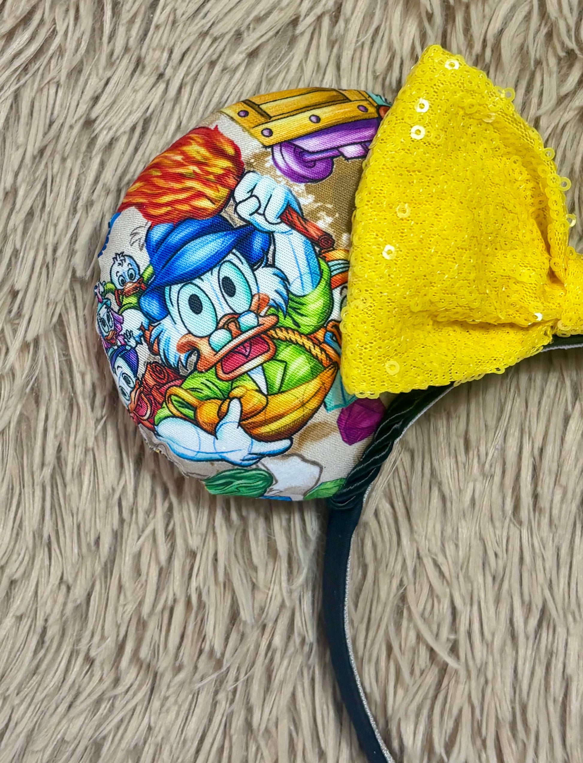 A close-up of headband featuring a colorful cartoon duck character print and a large yellow sequined bow, laid flat on a beige textured faux fur background.