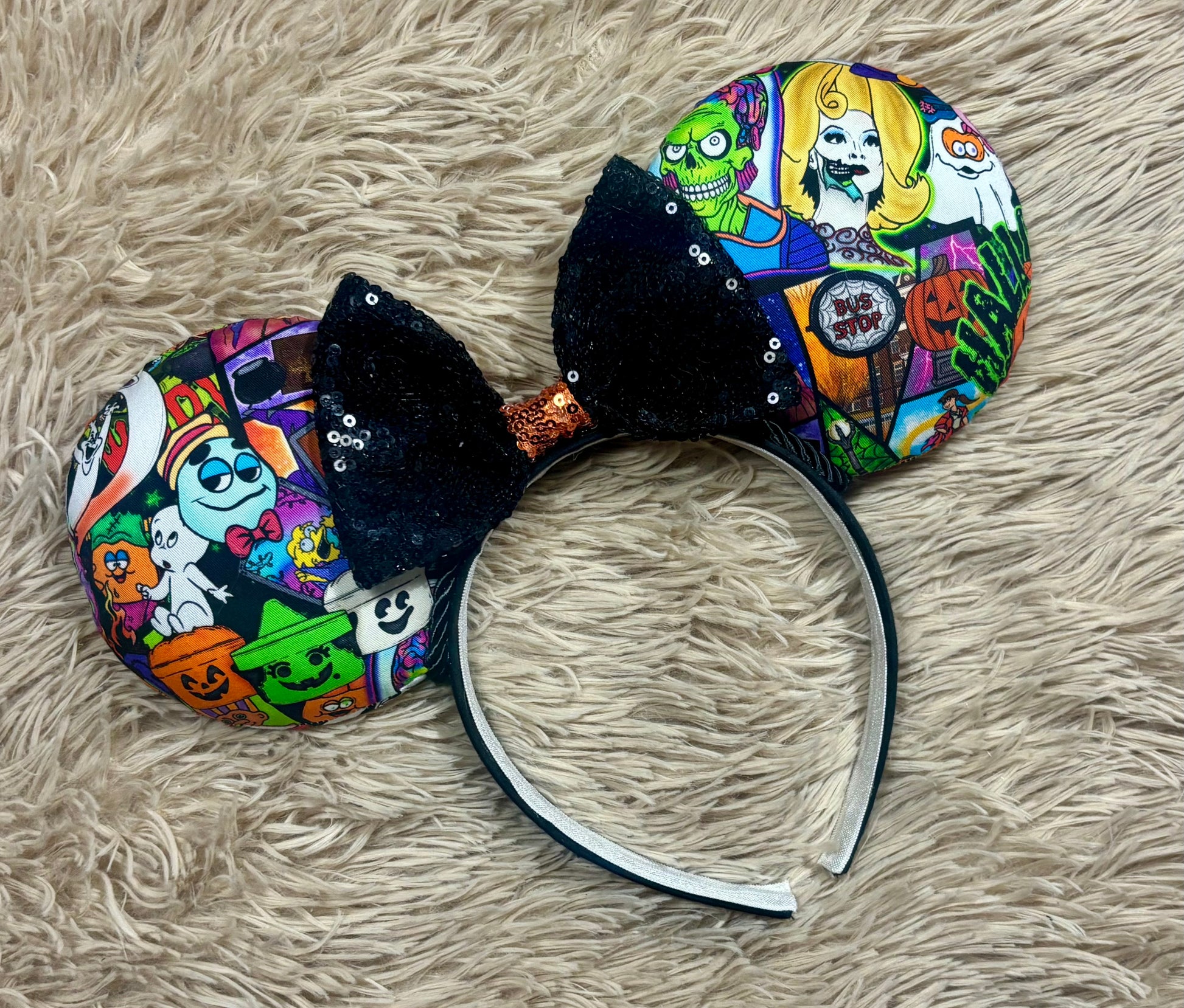 An ear headband accessory featuring a colorful Halloween-themed print on the outer ears and a black glitter bow in the center with black twisted trim along the inner ears.