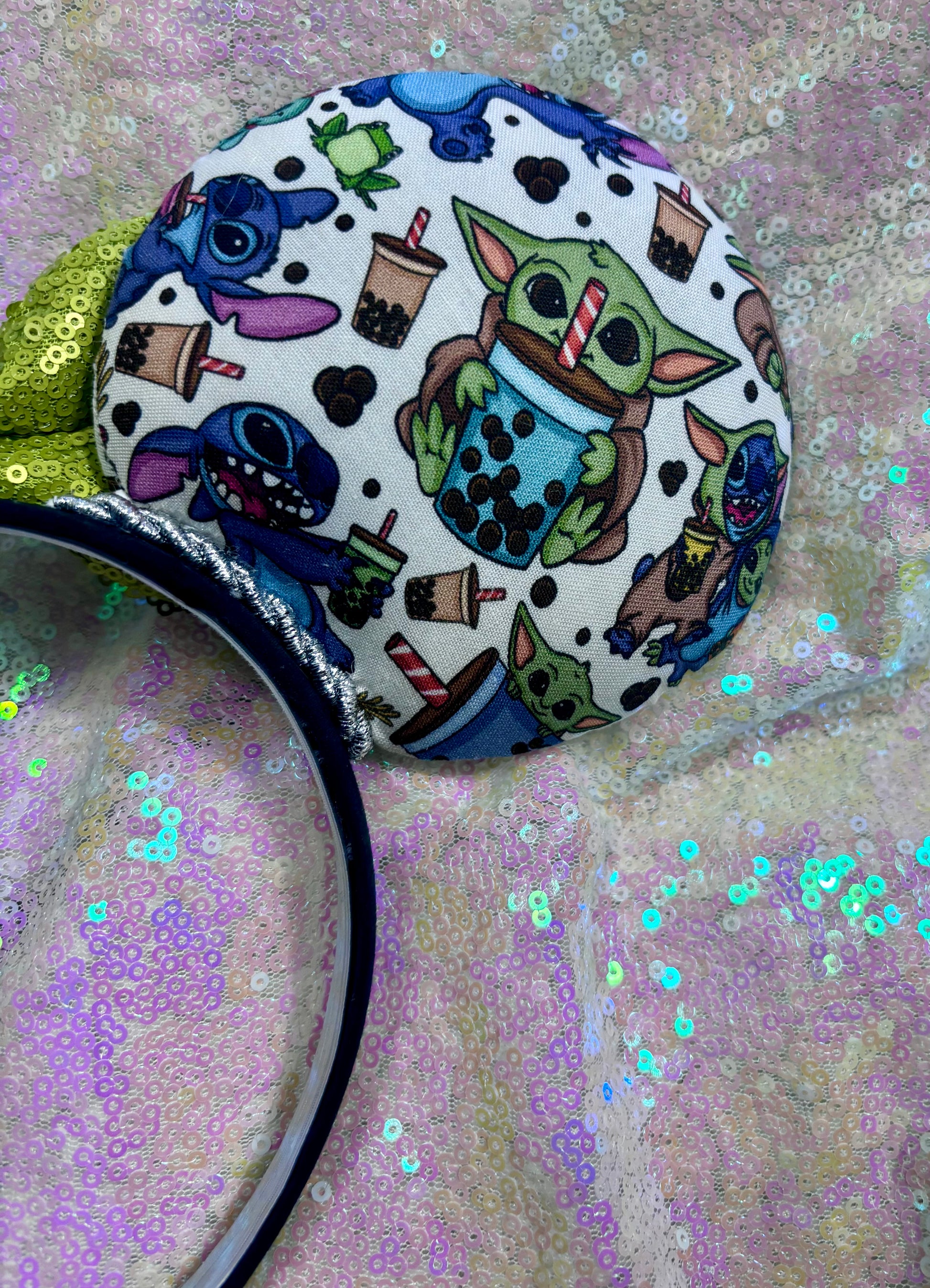 Handmade and ready to ship. Stitch and Baby Yoda are drinking coffee on the front and back with a lime green sequin bow. Back right mouse ear view. Shown on a white glitter background.