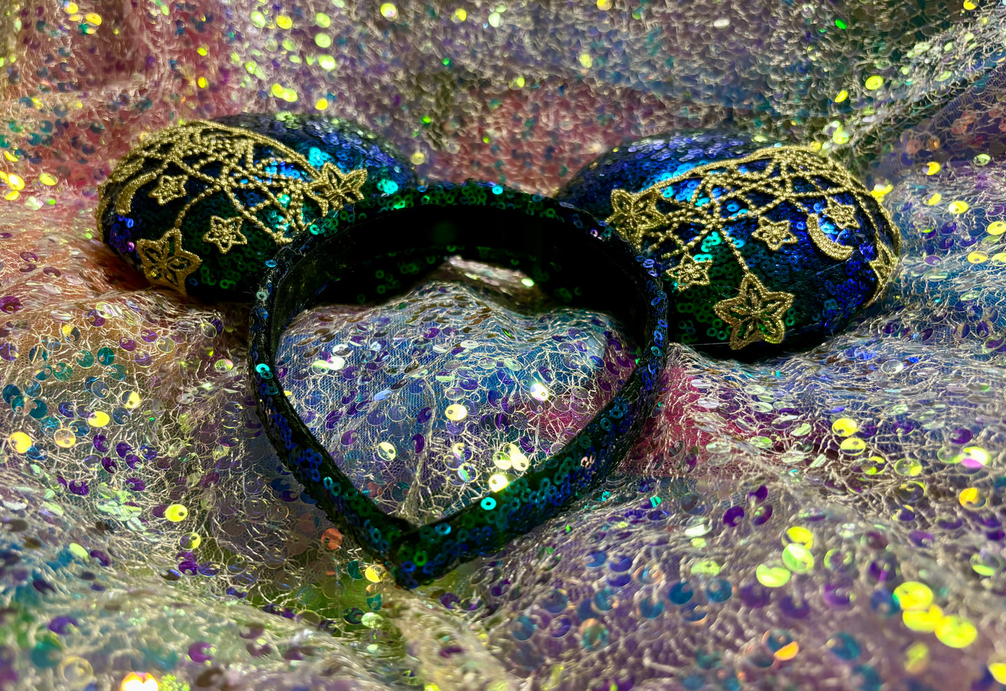 Mouse Ear headband that is a Handmade Iridescent blue and green sequins with gold sun, moon and stars attached to them. bow with star and moon patterns on a glittery background.