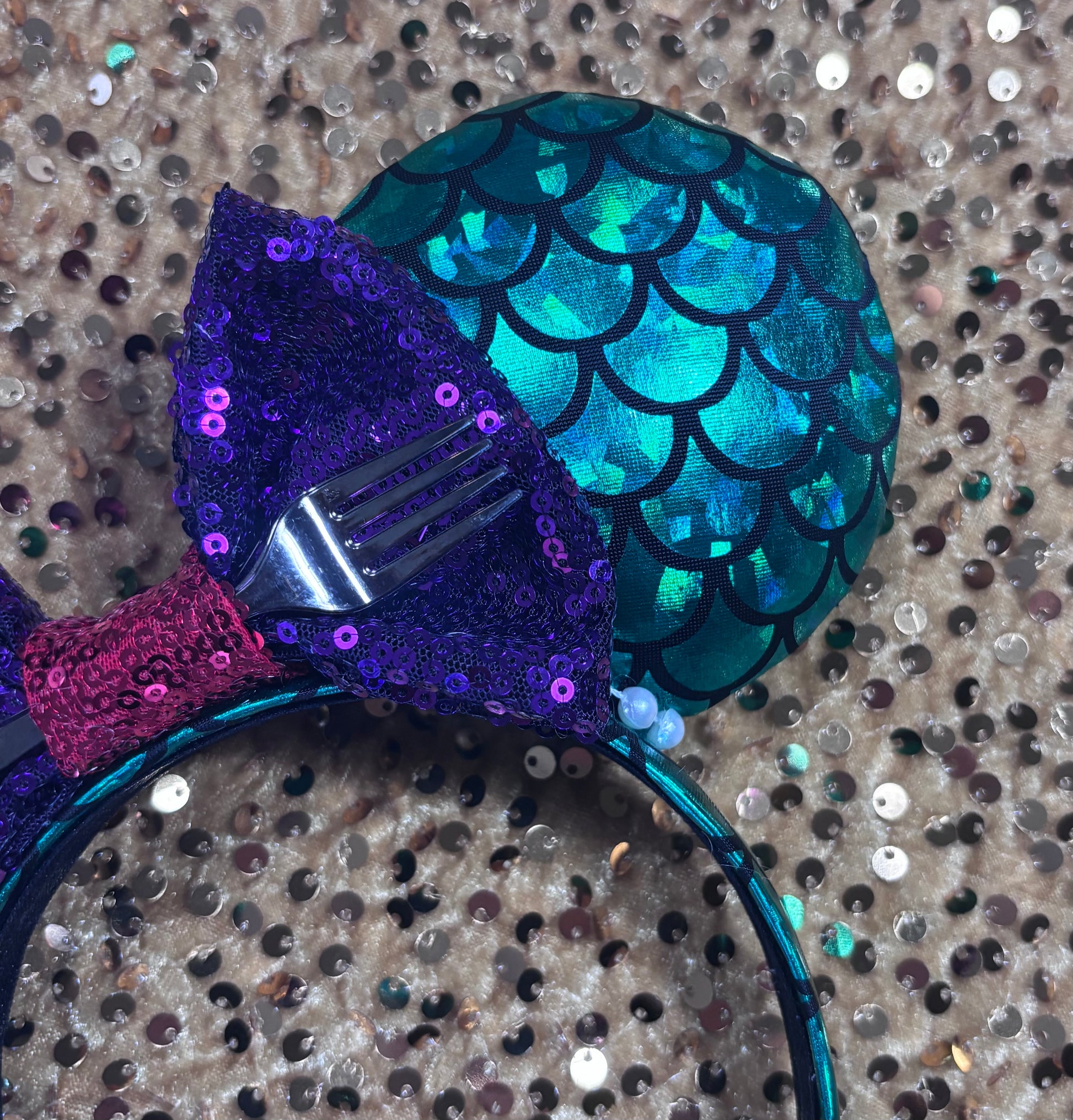 Mermaid-themed headband with teal scales and a purple sequin bow with a red middle and a dinglehopper middle piece. Pearl garland trim along inner ears m on a matching mermaid scales headband.