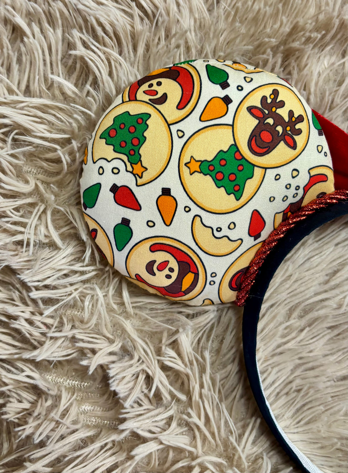 An ear headband featuring a festive Christmas print with red velvet bows, displayed on a beige faux fur background.