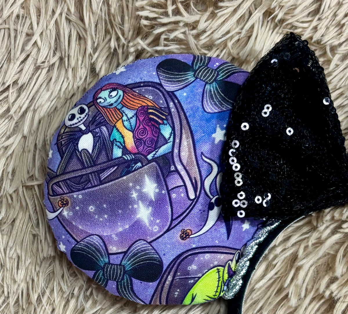 An ear headband accessory with a black sequined bow, featuring a purple HM and NBC themed print with a cartoon character in a buggy. Close up of front ear.