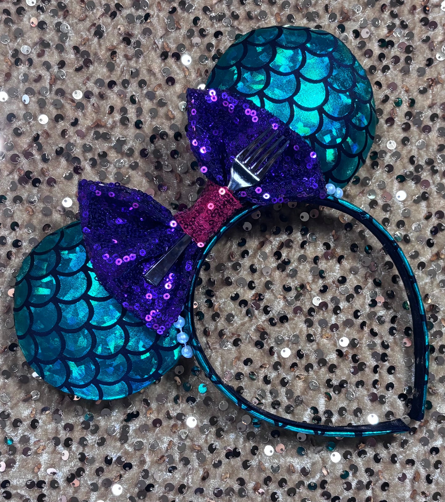 Mermaid-themed headband with teal scales and a purple sequin bow with a red middle and a dinglehopper middle piece. Pearl garland trim along inner ears m on a matching mermaid scales headband.